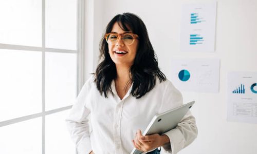 Data analyst standing in an office and looking at the camera. Portrait of a happy business woman holding a laptop in her hand.